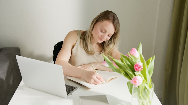 A woman sits at a desk writing in a notebook with a laptop and tulips nearby.