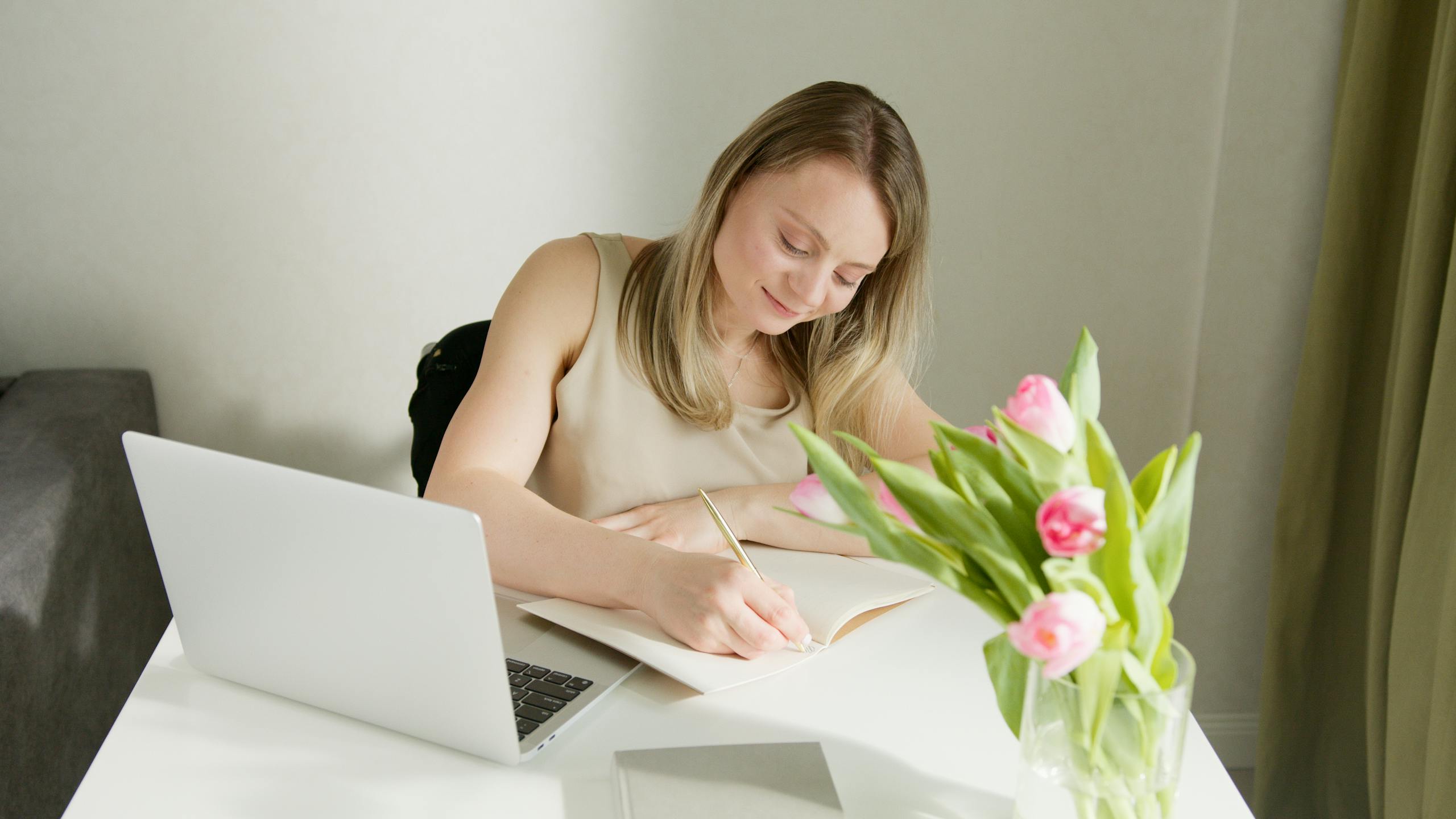 A woman sits at a desk writing in a notebook with a laptop and tulips nearby.