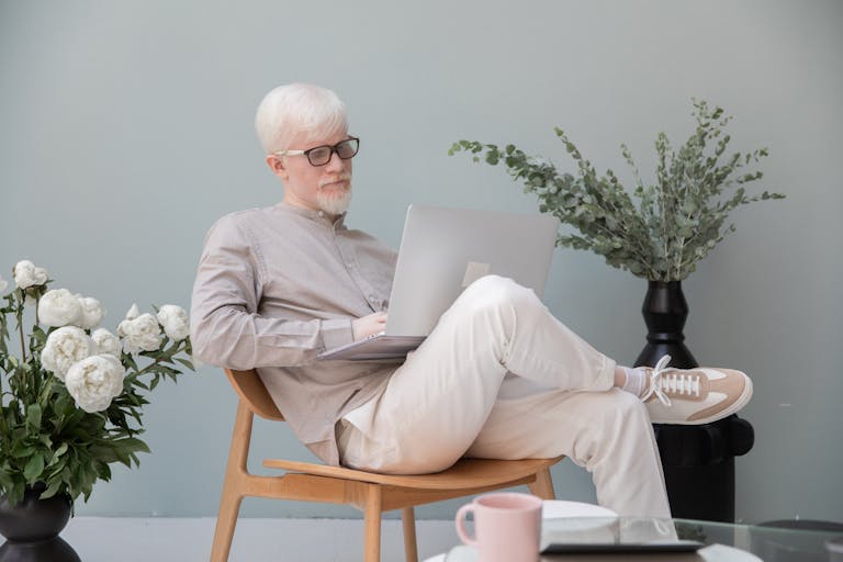 Concentrated male in eyeglasses with white hair and beard sitting in room near flowers while typing on computer during work
