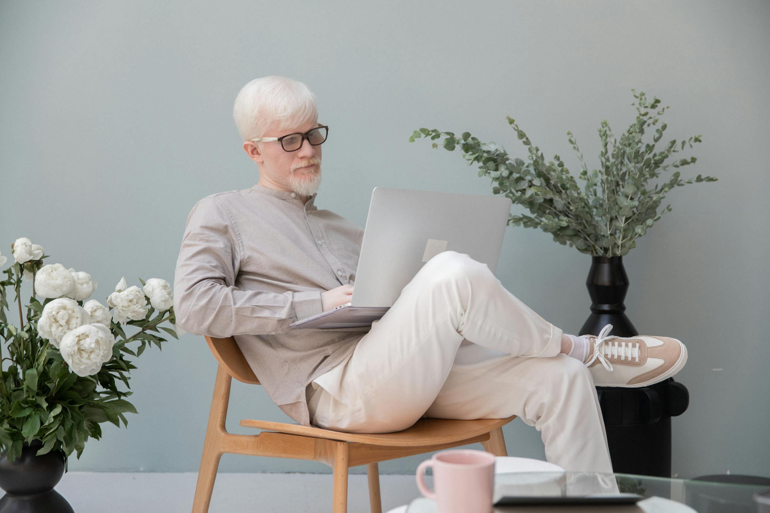 Concentrated male in eyeglasses with white hair and beard sitting in room near flowers while typing on computer during work
