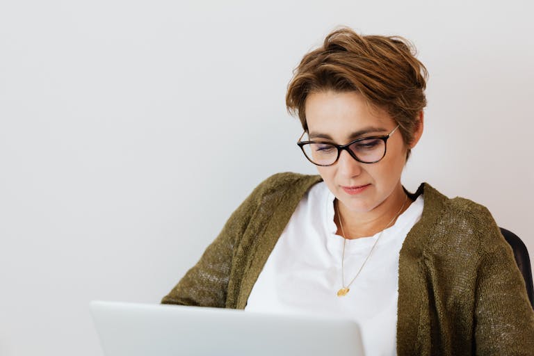 Smiling adult short haired female in stylish clothes and eyeglasses using laptop in light office space